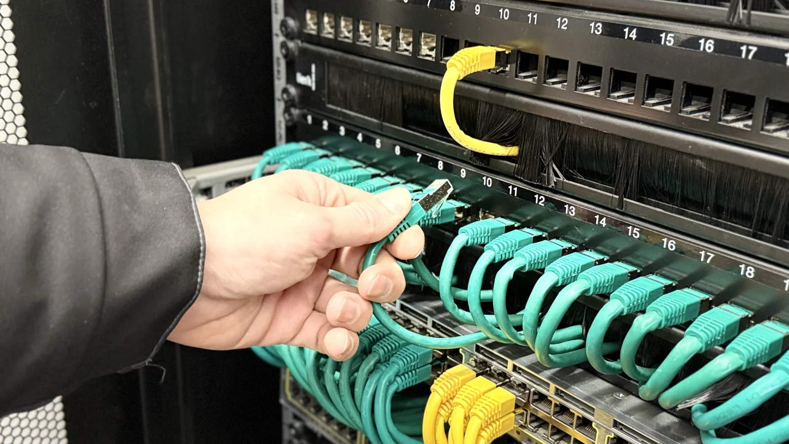 Une main tient une fiche RJ45 verte devant les ports et cordons d’une baie réseau.